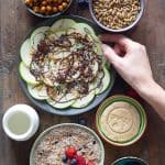 Cereal in a bowl, cookie on a plate, crispy chickpeas in a bowl, seeds in a bowl, all on a wooden table.
