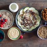 Cereal in a bowl, cookie on a plate, crispy chickpeas in a bowl, seeds in a bowl, all on a wooden table.