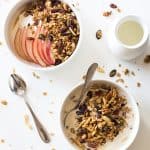 Healthy Homemade Granola in a bowl with milk and a spoon, and topping another bowl of yogurt and sliced apple. A spoon and milk jar next to it.