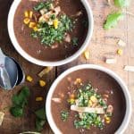 Two bowls of Mexican Black Bean Soup on a wooden board, two spoons and a small bowl of tortilla strips.