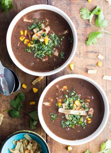 Two bowls of Mexican Black Bean Soup on a wooden board, two spoons and a small bowl of tortilla strips.