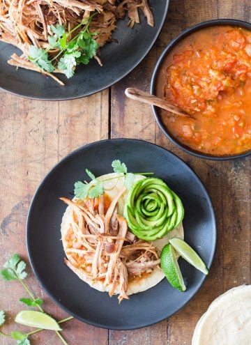 Top view of a pulled pork taco, a bowl of red salsa with a wooden spoon, and part of a plate with pulled pork.
