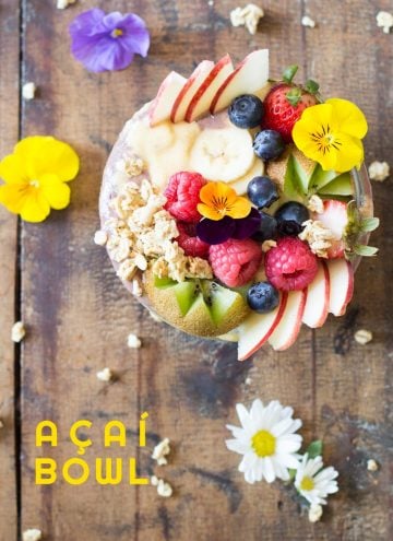 Top view of Acai Bowl topped with fresh fruit and flowers, on a rustic wood table with flowers, and text overlay.
