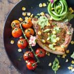 Mexican Pork Chops garnished with avocado cilantro sauce, corn kernels, cherry tomatoes and sliced lemon on a black plate.