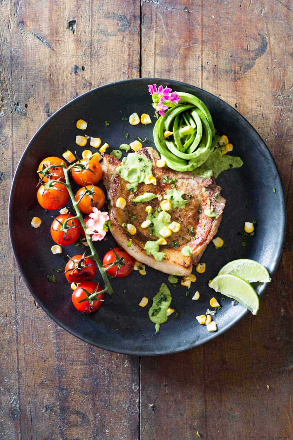 Mexican Pork Chops garnished with avocado cilantro sauce, corn kernels, cherry tomatoes and sliced lemon on a black plate.