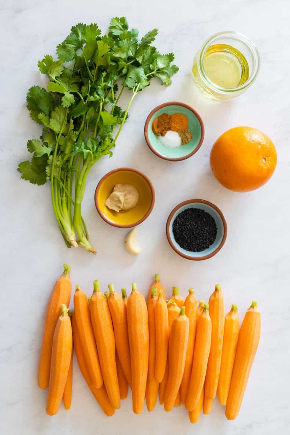 Carrots, cilantro, orange, oil, spices, garlic, sesame seeds laid out on a kitchen counter.