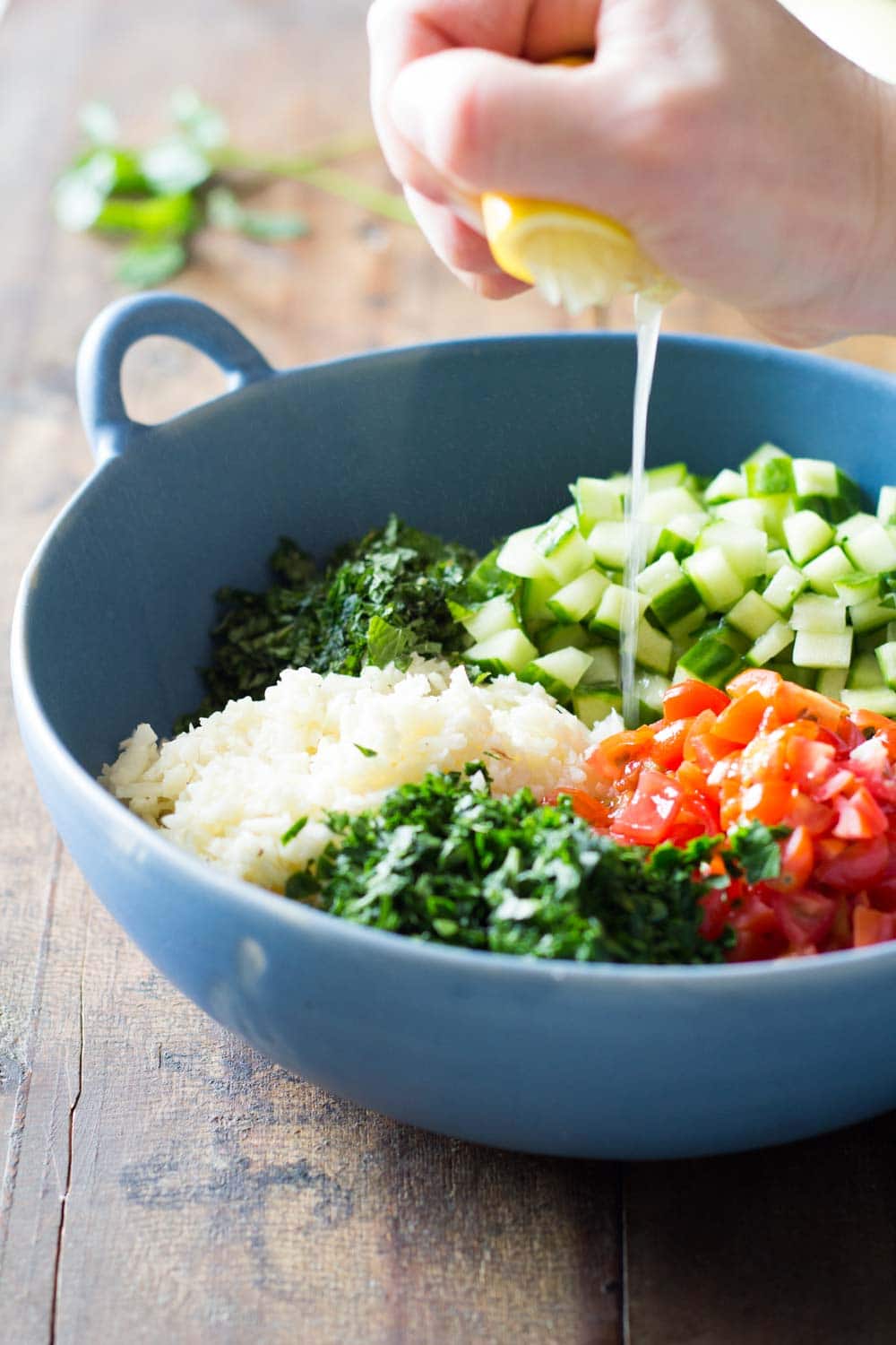 Cauliflower couscous with chopped parsley, diced cucumber and tomatoes in a blue bowl, with a hand squeezing a lime over it. 