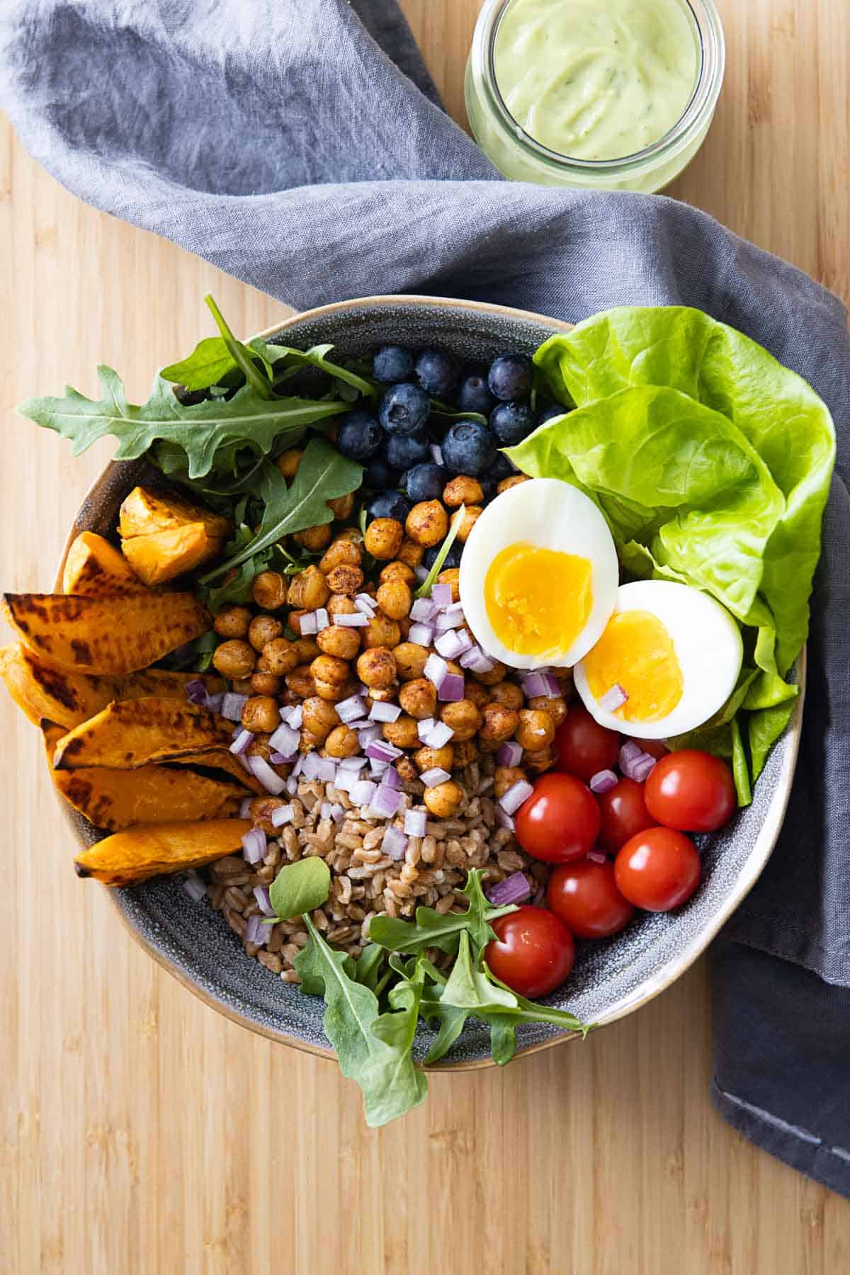 Bowl filled with lettuce, roasted sweet potato, roasted chickpeas, tomatoes, farro, red onion, blueberries, and a hard-boiled egg.