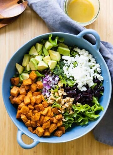 Lettuce, roasted sweet potato, avocado, onion, feta, cranberries in a blue salad bowl, dressing on the side.