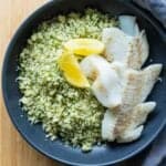 haddock on a bed of couscous with herbs and two lemon sliced on top.
