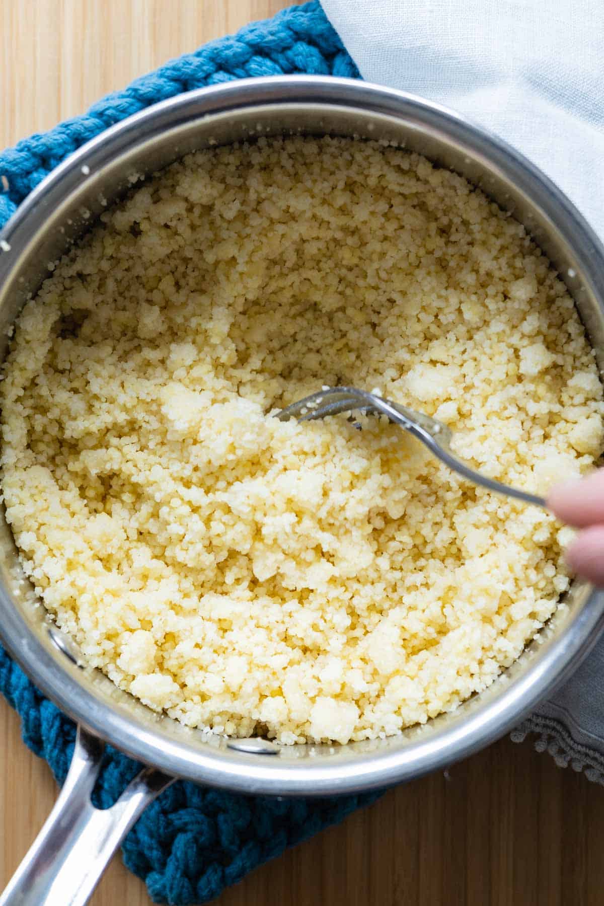 Stainless steel pot with cooked couscous and fork inside fluffing the couscous.