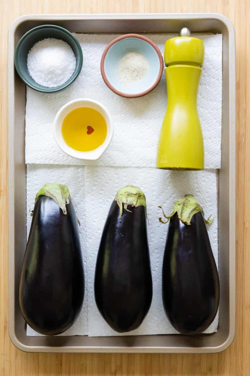 Eggplants, a bowl with oil, a bowl with salt, a bowl with herb salt, and a pepper mill laid out on a baking sheet.